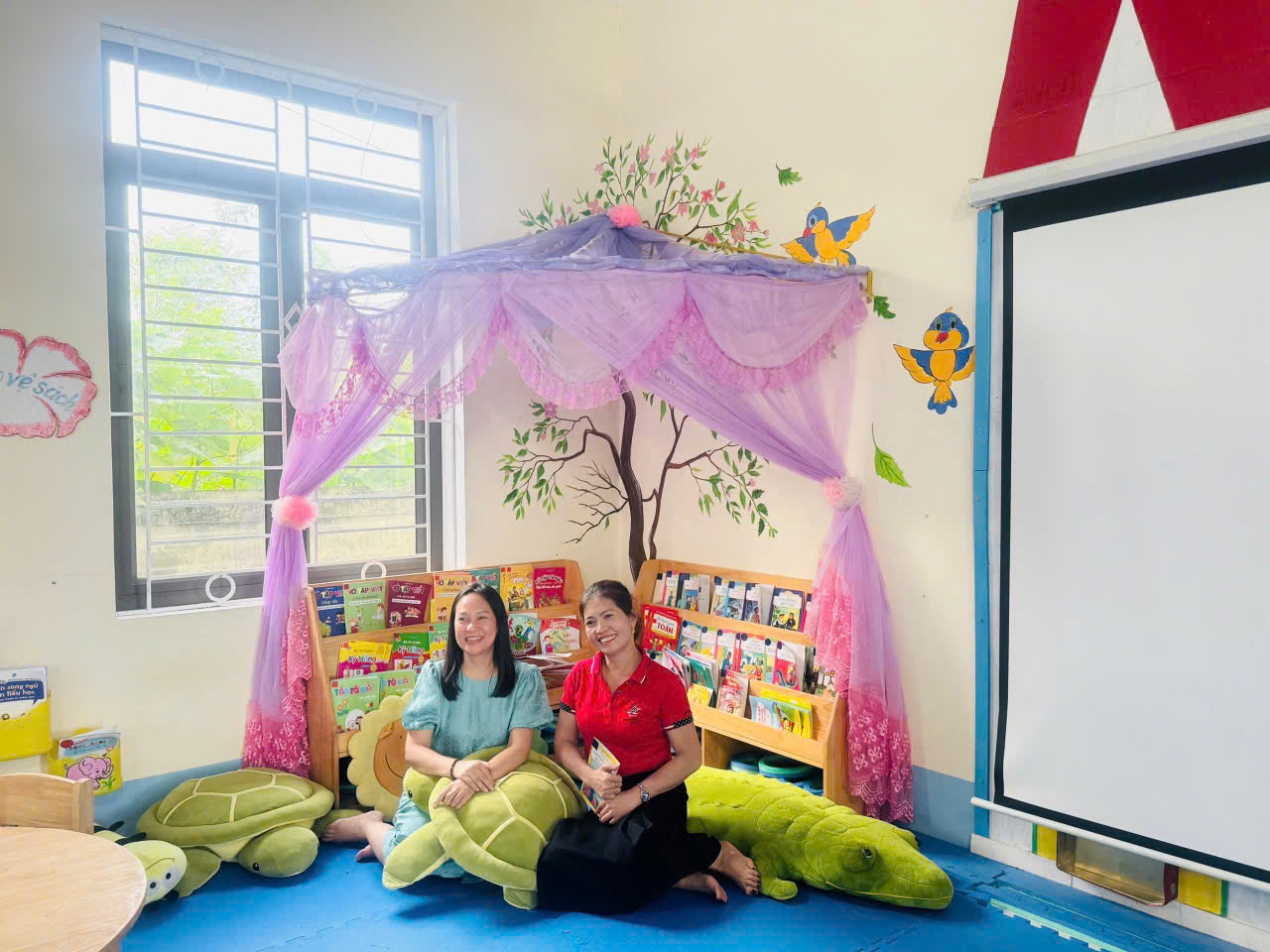 Góc đọc sau khi hoàn thiện: giá thấp ngang tầm trẻ, đệm ngồi, và tranh do chính các em vẽ lên tường.